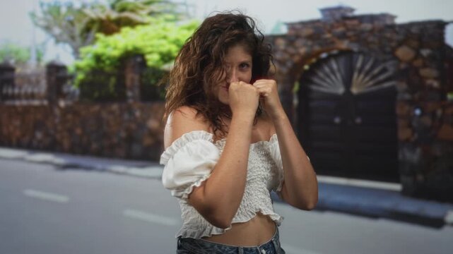 Woman with fists raised in boxing guard showing exposed shoulder and midriff on street by a house entrance; confident youthful fun.
