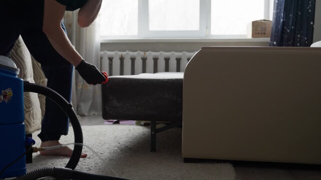 White Man Vacuuming Sofa Seams Vigorously, Moving Cushions, Extracting Dust And Debris, Lowlight Living Room, Blue Uniform, Black Gloves, Portable Extractor Unit Visible, Focus On Nozzle And Fabric
