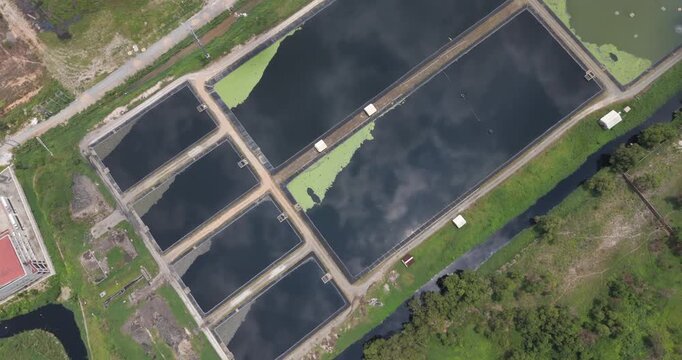 Top down aerial view of sewage treatment basins featuring dark water, green algae patches, and a bright sun reflection.