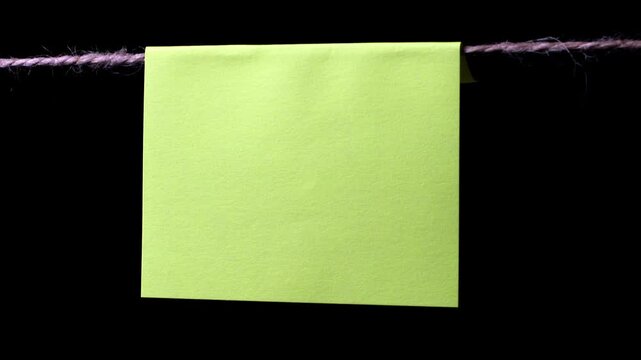 A square piece of yellow paper hangs on a thin rope against a black background. Space for text.