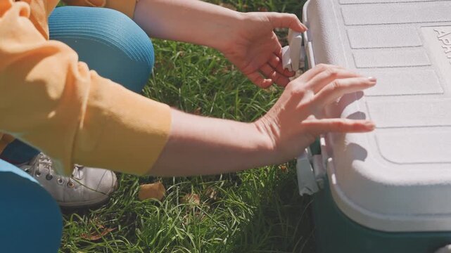 White camper tucks herbs into cooler, red cloth cushions produce while hands secure lid, practical meal prep on grassy campsite, readying salad and wrapped loaf for outdoor lunch.