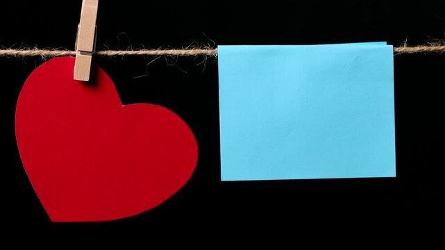A red heart and a square piece of blue paper hang from a thin rope against a black background, attached with a clothespin. Space for text.