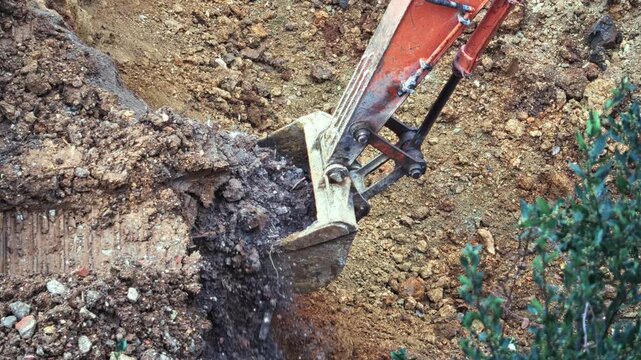 Excavator grading bucket digging a trench and dumping wet soil and rubble at a construction site, hydraulic arm and orange boom visible from above