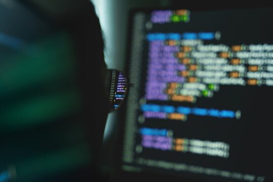 Finger taps keys beside glowing code on screen. Dark room highlights focused, intense concentration.