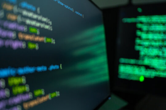 Dark room glows with green code on two screens.  Code scrolls in vibrant syntax, hinting at deep technical work