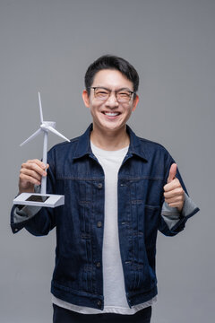 Smiling Asian man wearing glasses holding wind turbine model giving thumbs up isolated on gray background, ideal for renewable energy, ecology, and sustainability concepts.