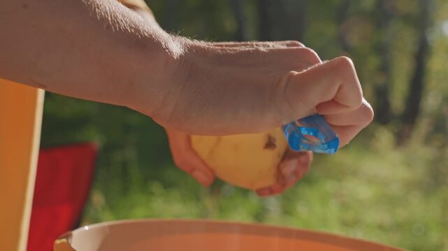 Closeup hands peeling potato by hand at campsite, white forager preparing wild tuber with blue peeler, sunlit birch clearing, focused slow movements, rustic ingredient preparation for camp meal