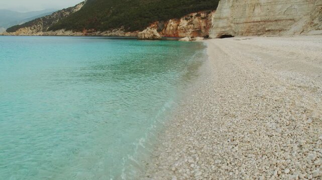 Crystal clear turquoise waves lapping pebble shoreline below green cliffs in Kefalonia Greece