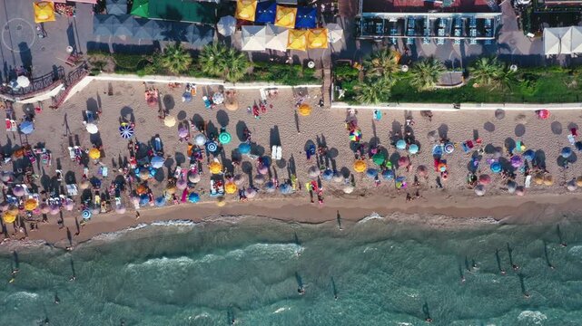 Aerial top-down view of crowded beach with rows of colorful umbrellas and swimmers entering the sea on a sunny summer day, Kusadasi, Turkey - May 01, 2025