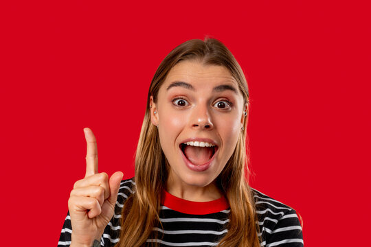 Happy woman having idea eureka moment, pointing finger up on red background. Clever lady showing answer gesture or remembered what she forgot, memory concept.