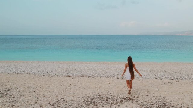 Young woman walking toward tranquil turquoise sea on empty pebble beach
