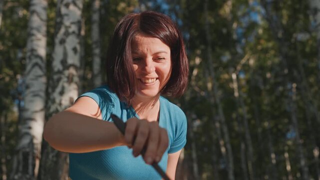 White woman camping stirring pan near birch trees. As camper, trail cook, forager, forest guide, and storyteller she smiles while pouring from small kettle and tending portable stove, closeup hands,