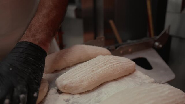 Baker shaping dough into loaves with gloved hands on a floured surface in a kitchen, showcasing the process of preparing fresh bread for baking