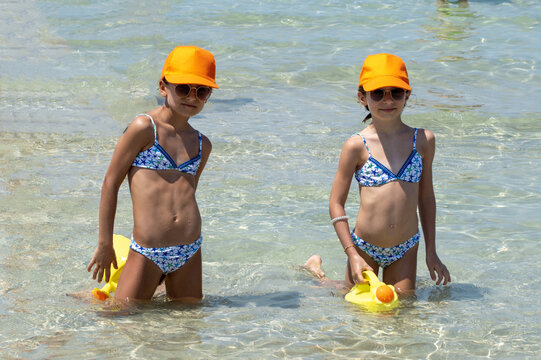 Sorelle gemelle di nove anni al mare con cappellino giallo mentre giocano con l'acqua.