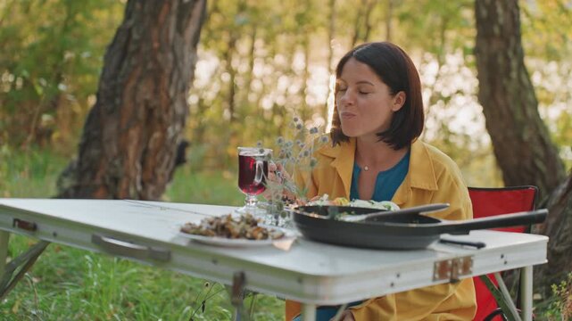 Caucasian woman camping blowing hot food weekend camper cooling steam from skillet with playful expression, birch trees and dusk glow, folding table with glass of red beverage, cozy outdoor solitude