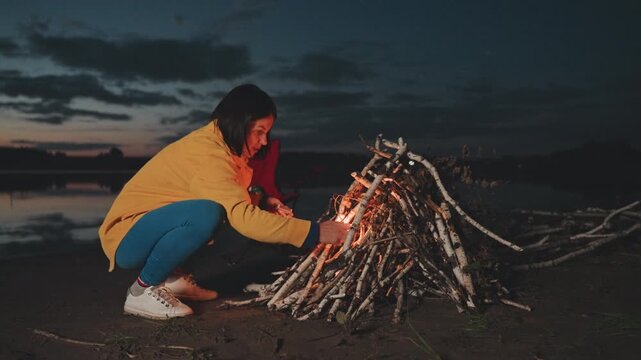 Young white woman lighting beach campfire wearing yellow jacket and jeans, squatting by driftwood teepee at dusk at lakeshore, sparks from lighter ignite kindling, warm glow contrasts cool twilight,