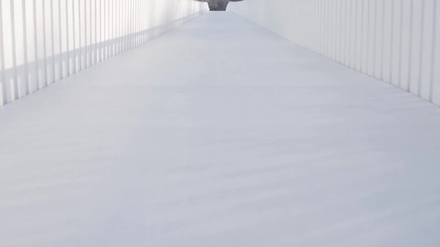 Tracking shot moving forward through empty white enclosed walkway between vertical panel walls on elevated pedestrian bridge with linear shadows and vanishing perspective