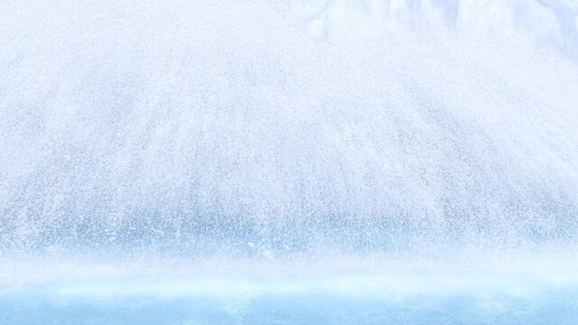 Slow motion pan tracking of waterfall mist and spray cascading into icy lake shore with falling snow and translucent blue foam in wintertime closeup panoramic view