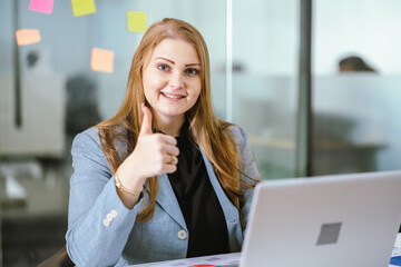 Obraz premium Smiling businesswoman blonde hair blue eyes formal suit sitting at desk giving thumbs up positive emotion modern office laptop glass wall teamwork motivation success