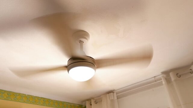 Motion blur of a ceiling fan with integrated light fixture rotating on a warm-toned ceiling. Cozy home interior with curtains and decorative wall border visible, suggesting comfort and airflow in a do