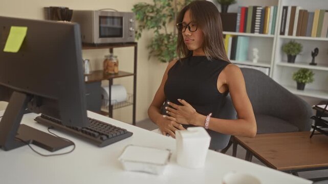 Thai woman clutching stomach at office building desk computer, leaning back in chair for stomachache; discomfort work.