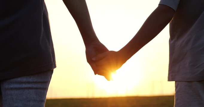 Close up shows mother and son holding hands against sun. Sun flare illuminates fingers holding hands. Trust and love appear in gesture of holding hands at sunset.