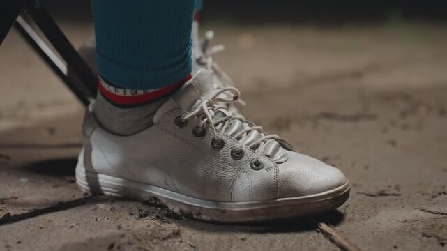 Closeup silver sneaker on camping ground, muddy laces and teal sock visible, folding chair leg supporting relaxed camper, subtle movement as foot presses into soil, evening light highlights texture