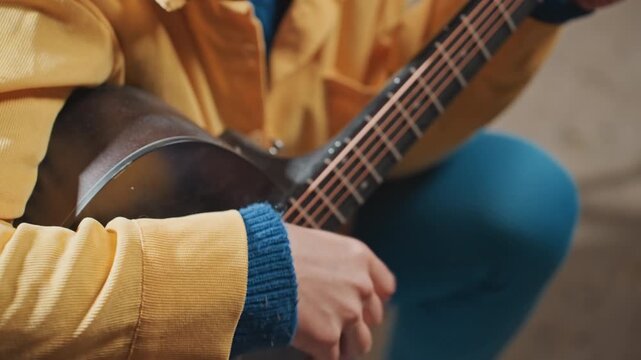 Closeup hands strumming acoustic guitar, camping vibe. Yellow jacket and blue leggings frame tight shot of fretboard and strings. Finger motion and textured sweater cuff create tactile intimate