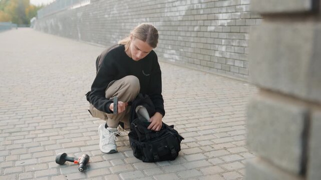 White photographer crouching by backpack remote, checking drone controller and gimbal, compact camera gear on pavement, brick wall backdrop, focused setup for shoot, content creation energy