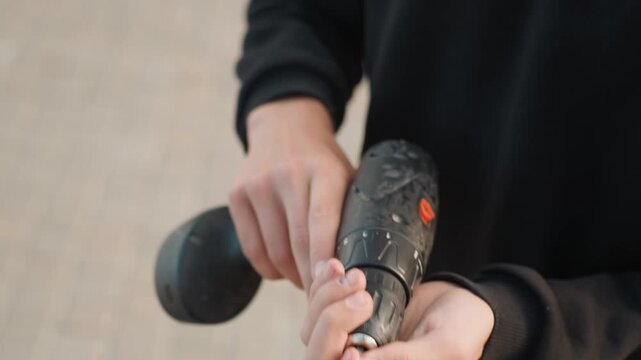 Closeup hands holding drill outdoors, urban tiled ground, black sweatshirt, inserting bit into chuck, careful adjustment and inspection, slow deliberate motions, soft natural light, tutorial vibe