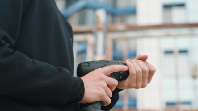 Closeup hands holding cordless drill prepping bit against scaffolded building exterior, focused grip and finger on trigger, battery indicator visible, urban renovation atmosphere, handson builder