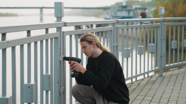 Man pausing beside rusted railing holding drill, seated on riverside promenade, contemplative gaze toward water, relaxed posture and toolbox nearby, autumn light and urban solitude after maintenance