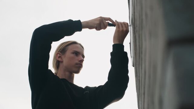 Man repairing concrete wall with screwdriver, closeup profile of installer securing metal bracket on urban facade under overcast sky, focused expression, dark sweater, precise hand movement, rooftop