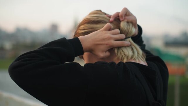 White man tying ponytail rooftop dusk, hands pulling blond hair into tight band, focused preparation for run or shift, concentrated posture with urban skyline backdrop, gritty yet calm mood, cinematic