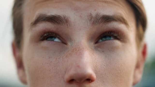 Close caucasian teenage male eyes glancing sideways, curious candid expression with subtle squint and blink, freckled skin in soft evening light, outdoor background suggesting alert observation.