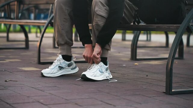 Person tying white sneakers on bench, closeup of hands working laces, white athletic shoes on textured pavement, cargo pants, metal bench frame, urban park atmosphere, soft morning light, fleeting