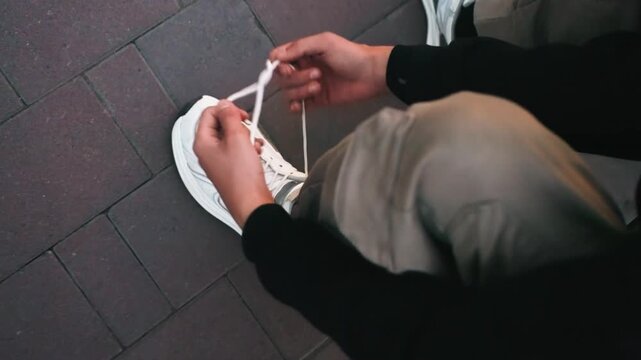 Overhead closeup tying white sneaker laces showing hands adjusting knot on pavement, casual outfit and focused posture, candid street routine before commute, soft morning light, textured sole and long