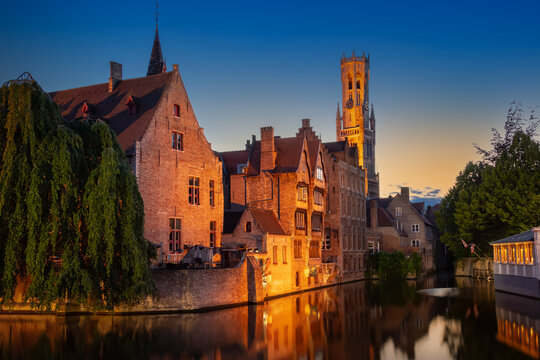 Canal view of Rozenhoedkaai and the Belfry of Bruges at dusk in Belgium. Historic medieval architecture reflects in the Dijver canal water under a twilight sky in the Flanders region.