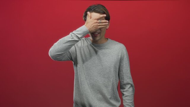 Man covers eye with hand showing ring and earring in studio red backdrop, smiling while peeking then closing face; amusement.