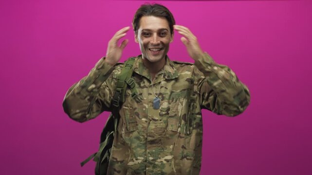 Soldier man in camouflage uniform with dogtags and backpack straps raises open hands in studio against magenta backdrop; surprise doubt.