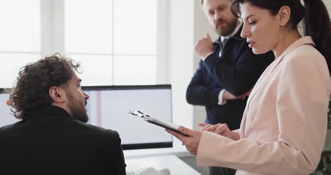 Team colleagues and managers in office meeting. Woman leader holds clipboard, briefing coworkers at computer screens on tasks and planning during discussion. Teamwork and leadership made clear.