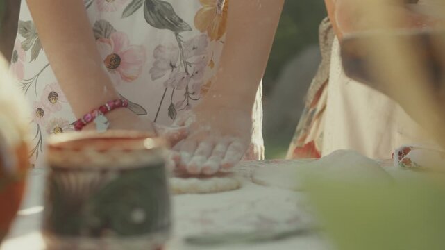 Young girl with floral dress and bracelet kneads dough with hands on table, surrounded by traditional pottery and kitchenware in a bright outdoor setting