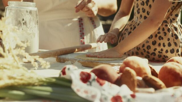 Young girl in leopard print dress rolls dough with a wooden rolling pin on a table surrounded by fresh fruits and vegetables in a sunny outdoor kitchen setting