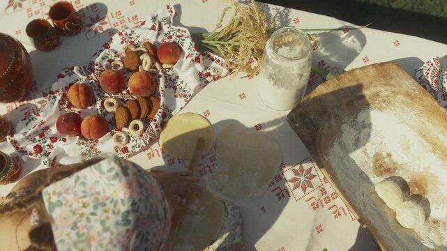 Female baker prepares dough on a decorated table with traditional pastries, flour, and ingredients, showcasing the process of making baked goods outdoors