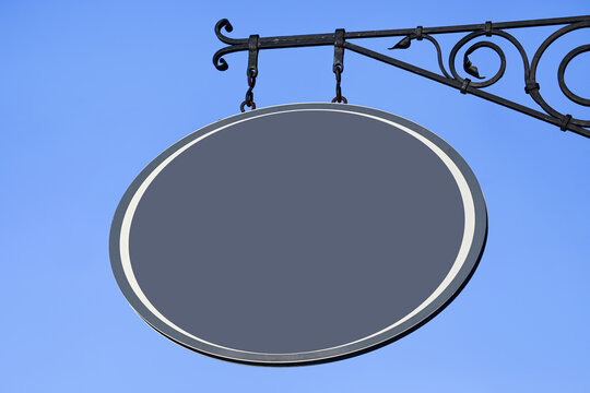 Blank oval hanging signboard on decorative metal bracket against blue sky. Empty business sign mockup for branding. Outdoor advertising space for shop logo and identity design.
