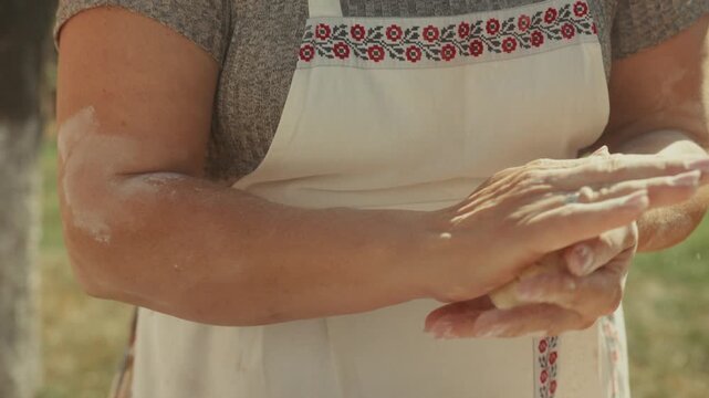 Woman with light skin in apron kneads dough with hands, shaping it on a table outdoors, surrounded by natural light and greenery in a rural setting