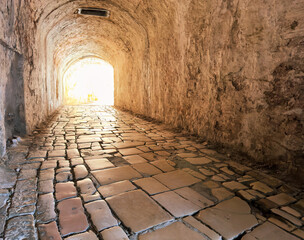 Fototapeta premium Stone tunnel passage inside the Old Fortress in Corfu Old Town, Greece. A bright exit over cobblestone path