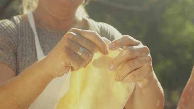 Woman preparing dough by stretching and shaping it into a thin round form outdoors, sunlight illuminating her hands and the soft texture of the dough