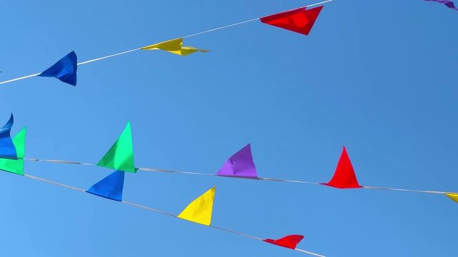 A string of colorful triangular flags hangs across a clear blue sky, waving in the wind. Red, yellow, green and blue banners create a festive mood.