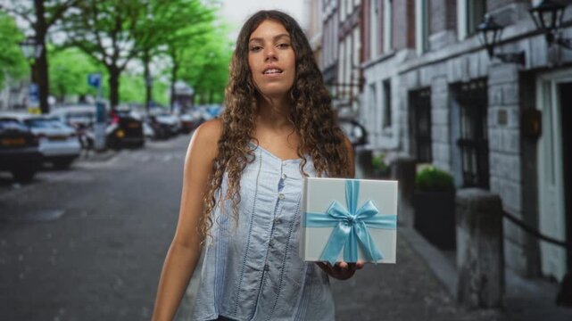Hispanic teenage woman holding gift box with blue ribbon, making ok sign to eye on street; celebration joy.